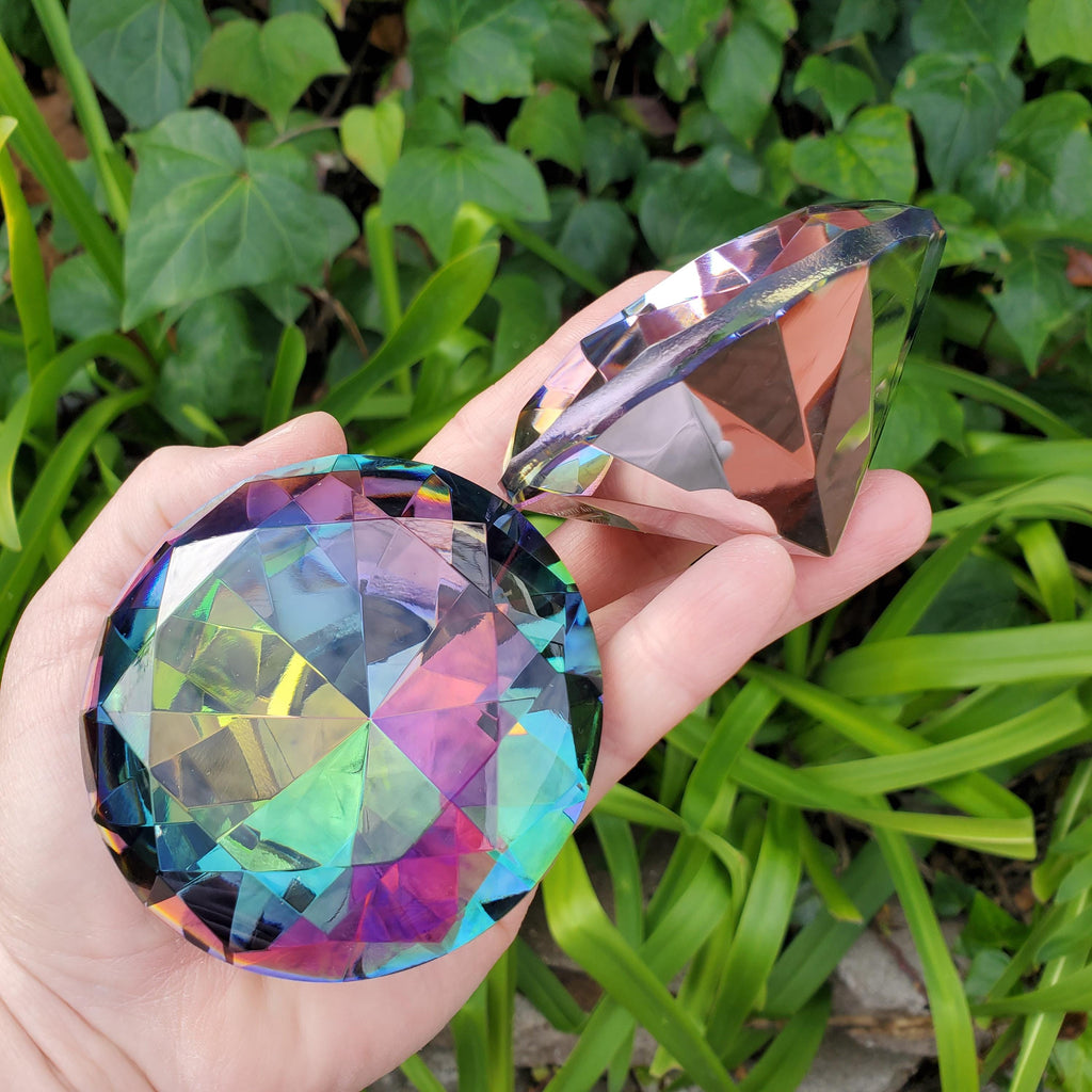 May include: Two iridescent glass diamond-shaped objects. One is a large, pointed-bottomed, faceted round shape. The other is a smaller, triangular, faceted shape. The objects reflect a rainbow of colors, including pink, green, and blue.