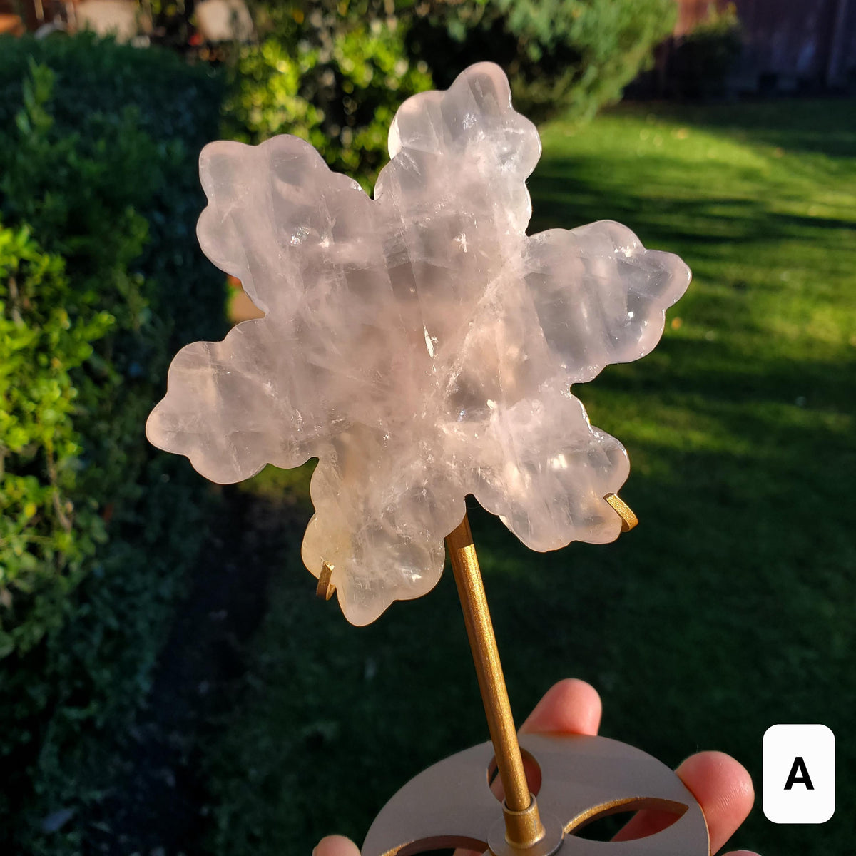 May include: A pink rose quartz crystal carved into a flower shape, mounted on a gold metal stand. The crystal is translucent and has a delicate, feminine appearance.
