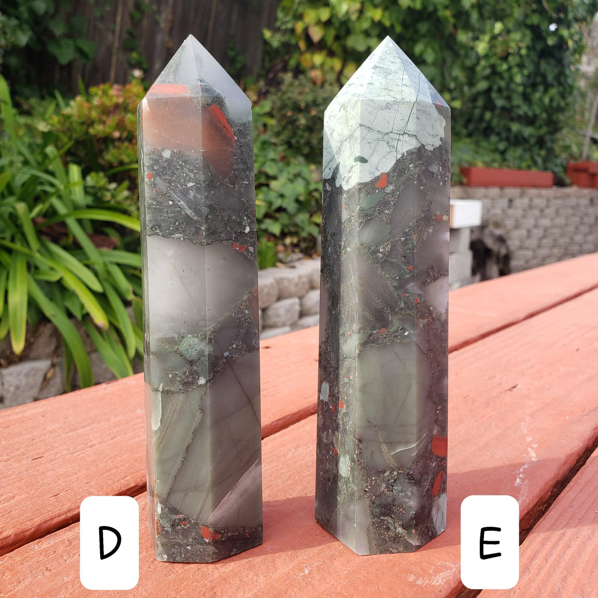 May include: Two green, red, and black crystal points, labeled D and E, are displayed on a wooden surface. The points are approximately 4 inches tall.