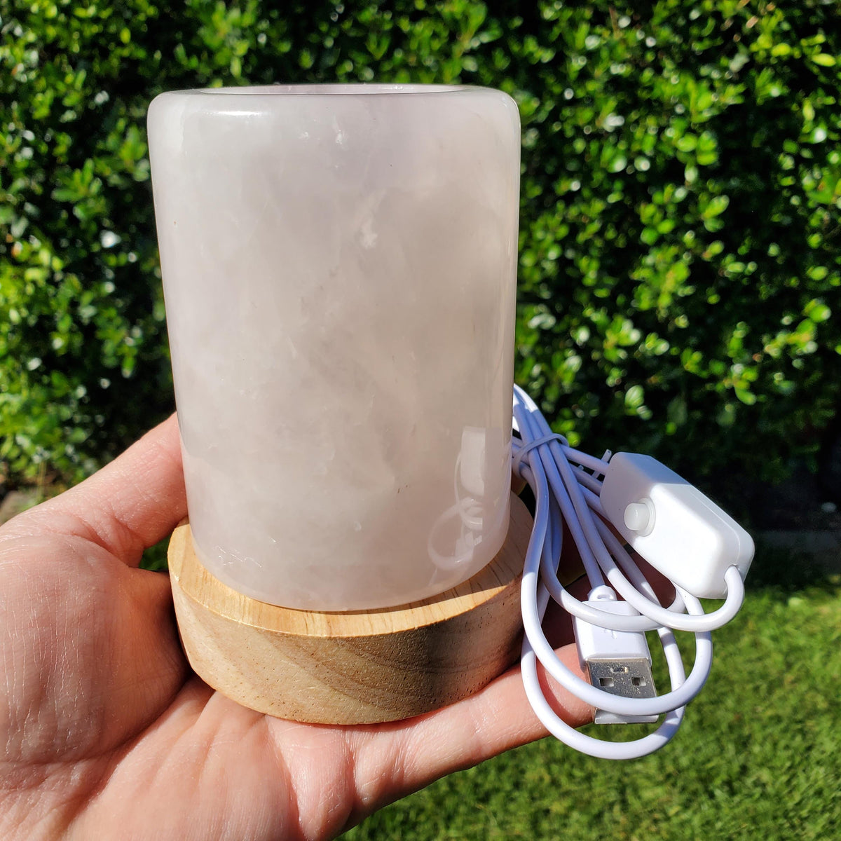 May include: A cylindrical, light pink rose quartz lamp sits on a round wooden base. A white USB cable with an inline switch extends from the lamp. The background is a blurred green hedge.