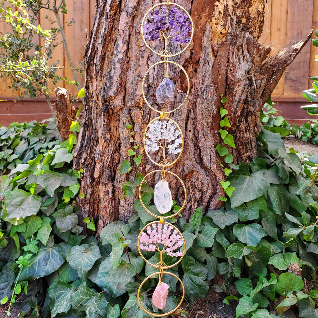 May include: A vertical hanging ornament featuring six gold-toned metal rings arranged in a column. The rings contain wire-wrapped tree sculptures made of purple, clear, and pink crystals, alternating with single raw crystal stones in purple, clear, and pink.