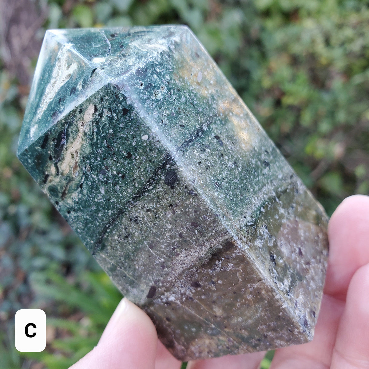 May include: A green, gray, and black crystal point with a rough, textured surface. The crystal is shaped like a pyramid with a pointed top.