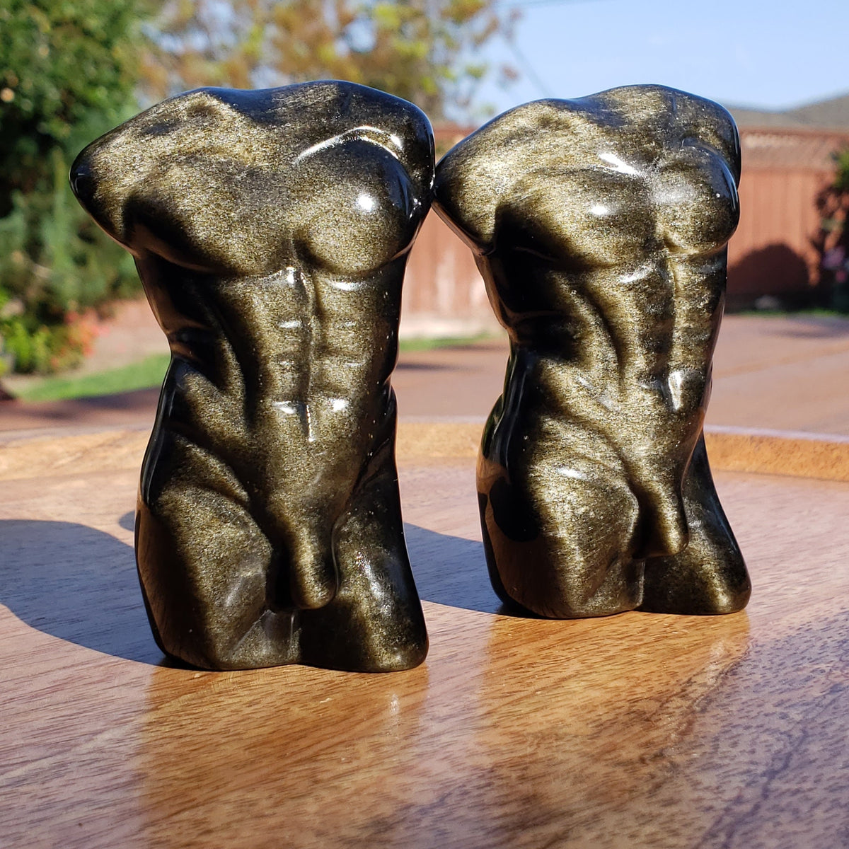 May include: Two polished golden obsidian sculptures depicting a human torso. Each piece features a smooth, reflective surface with a metallic gold sheen and dark black undertones, showcasing detailed abdominal muscles and a defined chest structure.